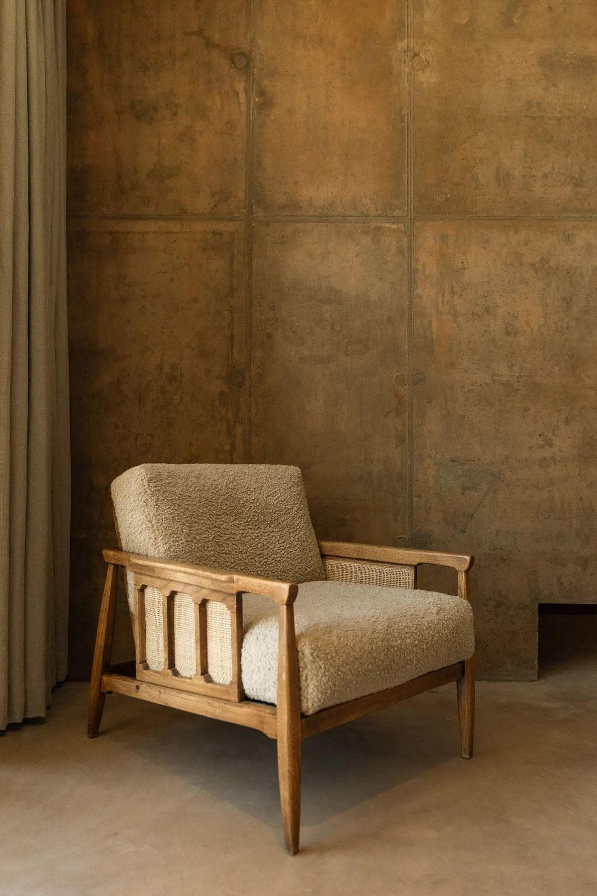 A bouclé-upholstered armchair with a solid wood and cane frame, set against a weathered rust-toned wall.
