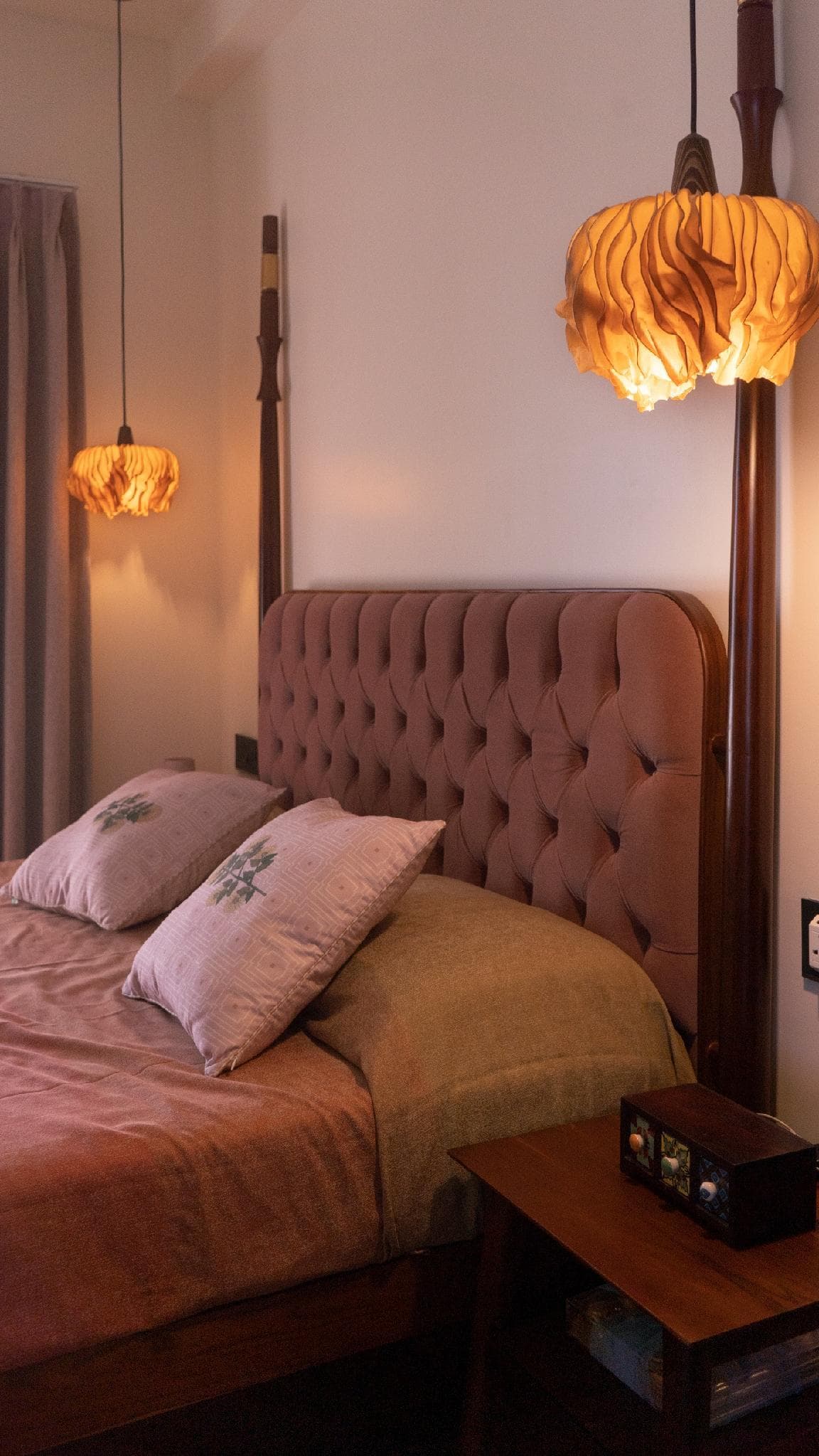 A four-poster bed with a tufted headboard, lit by two sculptural fabric pendant lights.