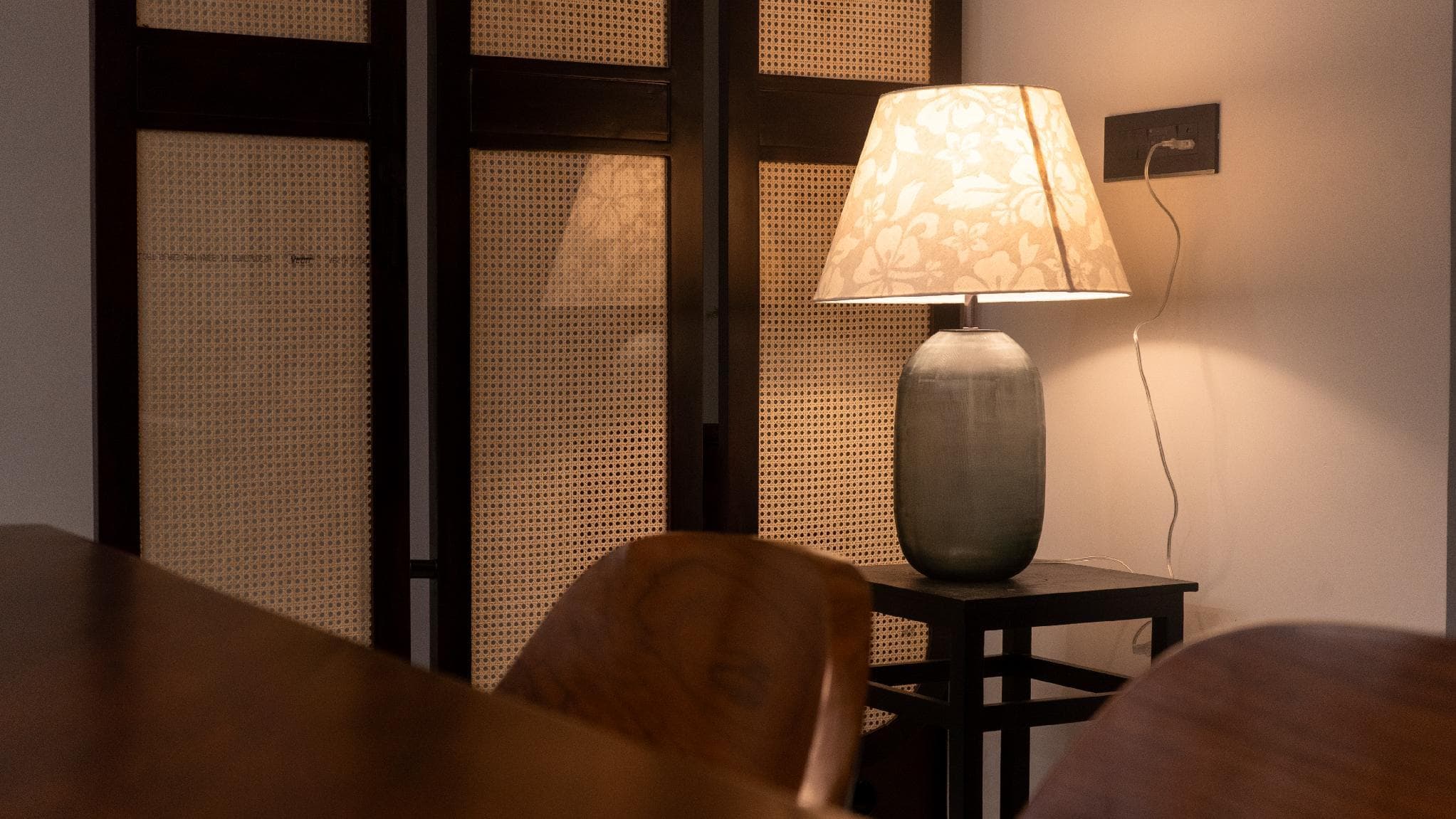 A ceramic table lamp with a floral shade beside a cane-panelled screen, warmly lit.
